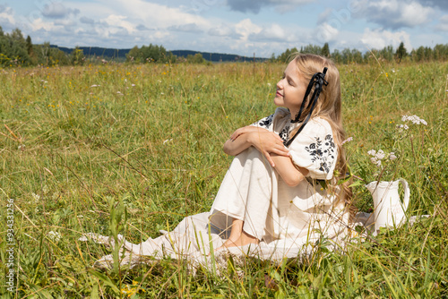 girl in white dress sits on grass and looks into the distance, in summer on meadow