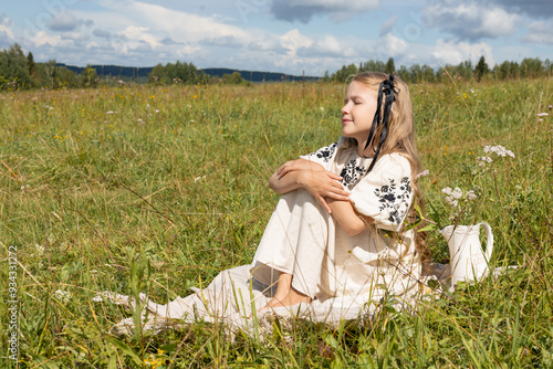 girl in white dress sits on grass and looks into the distance, in summer on meadow