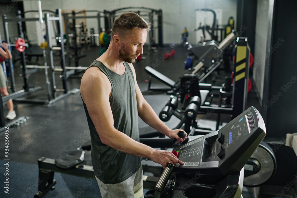 Caucasian man switching on treadmill to do cardio while his partner doing squats in blurred background Caucasian man switching on treadmill to do cardio while his partner doing squats in blurred background