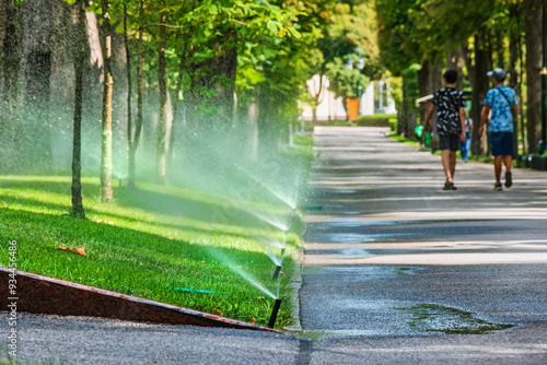 Automatic sprinkler system (irrigation system) for watering the lawn near the alley in the city park on a summer day. Flying splashes of water on the green grass photo