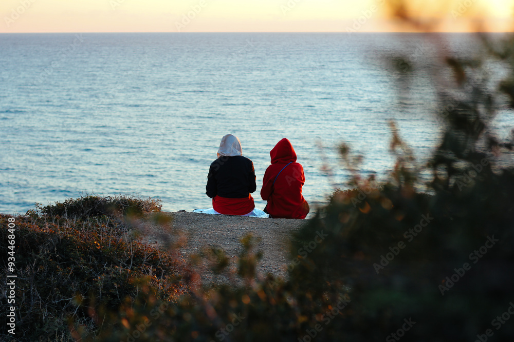 Close-up of two people sitting on a cliff overlooking the ocean at sunset. Sunset view, ocean horizon, outdoor relaxation, nature escape, calm moment, serene coastline, scenic landscape.