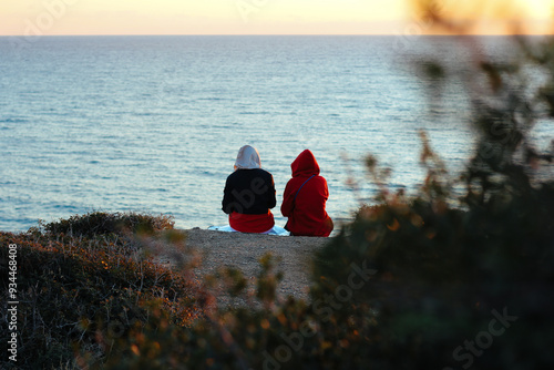 Close-up of two people sitting on a cliff overlooking the ocean at sunset. Sunset view, ocean horizon, outdoor relaxation, nature escape, calm moment, serene coastline, scenic landscape.