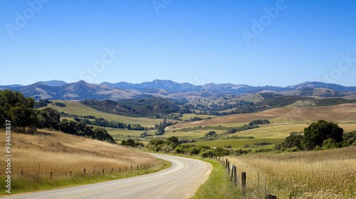 A tranquil landscape with a road curving through hills towards distant mountains, under a clear blue sky.