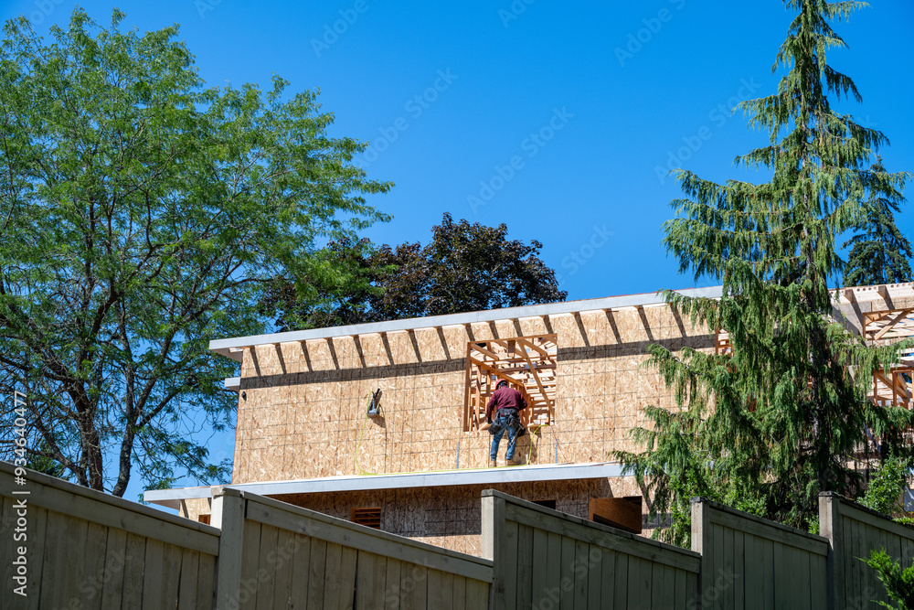View of new residential home construction over existing wood fence, worker on wall working in window opening of OSB oriented strand board exterior wall, summer construction season
View of new residential home construction over existing wood fence, worker on wall working in window opening of OSB oriented strand board exterior wall, summer construction season