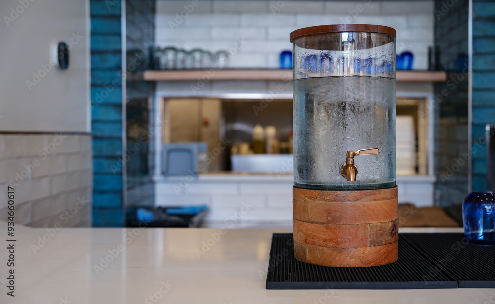 A wooden and glass water dispenser placed on a modern countertop setting in a kitchen or cafe environment, highlighting simplicity and function.