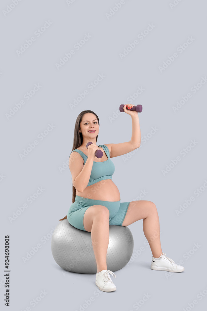 Fototapeta premium Sporty pregnant woman with dumbbells training on fitball against light background