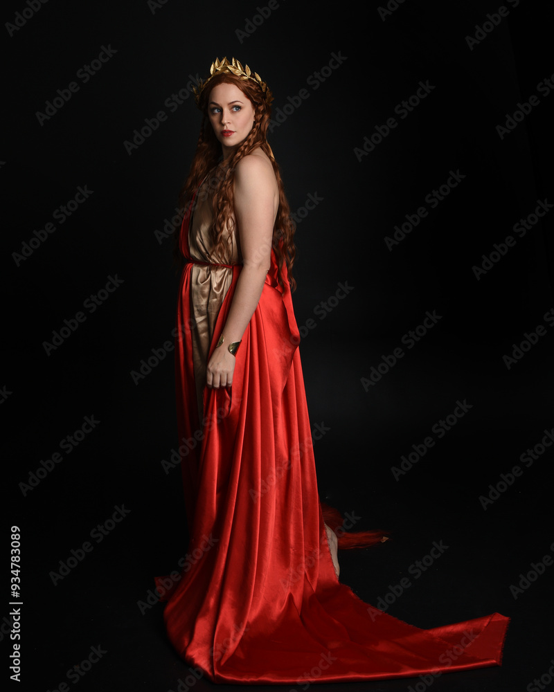 Full length portrait of beautiful female model wearing grecian red silk gold roman robes, historical fantasy goddess costume with crown wreath. Standing pose isolated on dark studio background. Full length portrait of beautiful female model wearing grecian red silk gold roman robes, historical fantasy goddess costume with crown wreath. Standing pose isolated on dark studio background.