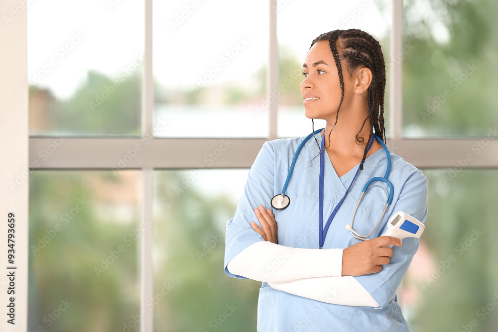Young African-American female doctor with stethoscope and thermometer near window in clinic Young African-American female doctor with stethoscope and thermometer near window in clinic