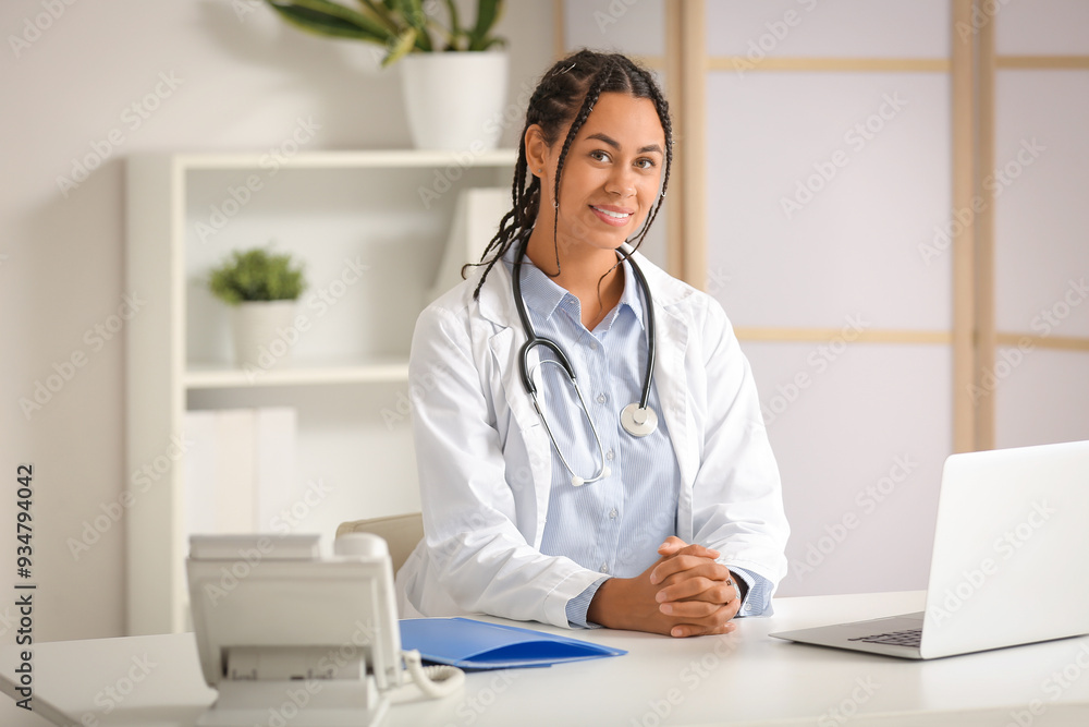 Young African-American female doctor with telephone and laptop sitting at table in clinic Young African-American female doctor with telephone and laptop sitting at table in clinic