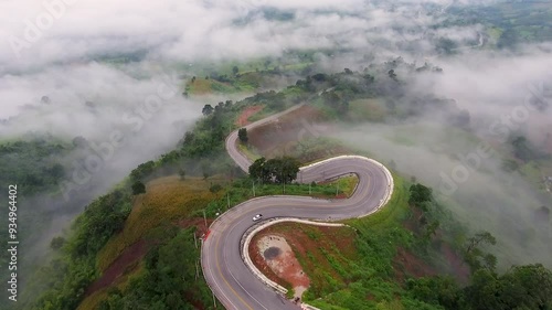Aerial view of winding road through green mountains, with a river flowing through the valley and a clear blue sky with fluffy clouds