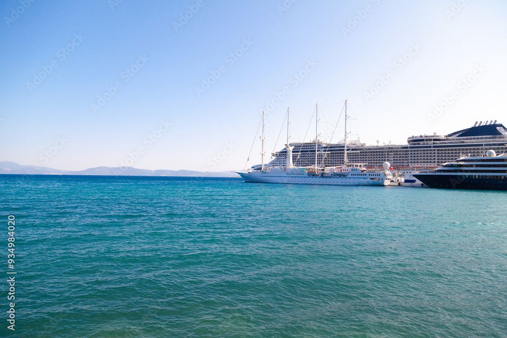 Obraz premium Large cruise ships and a sailboat are docked at the pier against the blue sea on a clear summer day. Ideal for content about maritime tourism and sailing.