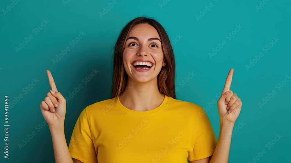 Joyful woman in yellow shirt smiling widely, pointing upwards against a vibrant teal background, exuding excitement and positivity in a cheerful atmosphere. Joyful woman in yellow shirt smiling widely, pointing upwards against a vibrant teal background, exuding excitement and positivity in a cheerful atmosphere.