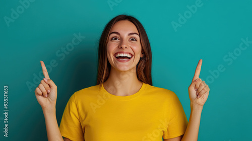 Joyful woman in yellow shirt smiling widely, pointing upwards against a vibrant teal background, exuding excitement and positivity in a cheerful atmosphere.