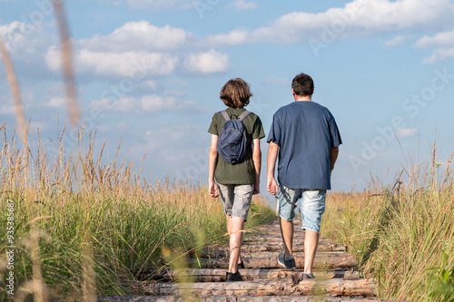 Father and his teenager son walking together on wooden road at field with tall grass, blue summer sky background, back view