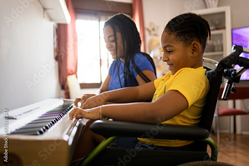 Two dark-skinned brothers are playing an electronic piano in their living room. The disabled child in a wheelchair is in the foreground.Concept of young africans playing a piano.