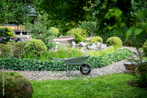 Wheelbarrow positioned on stone pathway surrounded by garden with hedge, plants, trees. Empty park with wheelbarrow in center. Landscape design, horticulture, plant maintenance, city greening. photo