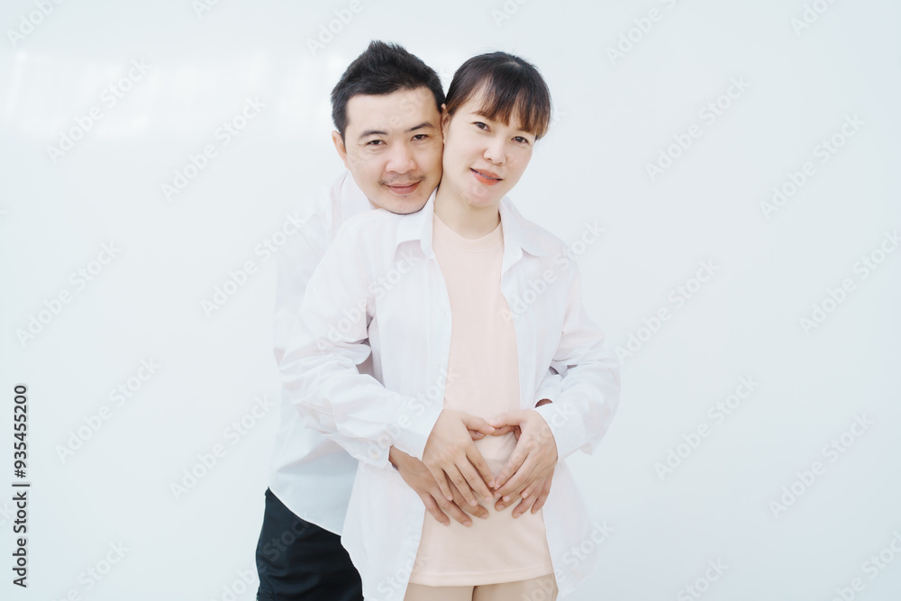 A happy Asian couple, expecting a baby, embraces in a cozy living room. The pregnant woman smiles warmly, while the man lovingly supports her, capturing a moment of joyful anticipation. A happy Asian couple, expecting a baby, embraces in a cozy living room. The pregnant woman smiles warmly, while the man lovingly supports her, capturing a moment of joyful anticipation.