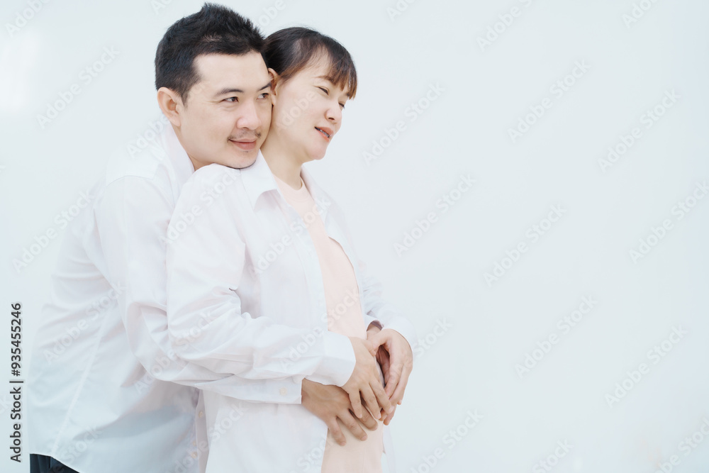 A happy Asian couple, expecting a baby, embraces in a cozy living room. The pregnant woman smiles warmly, while the man lovingly supports her, capturing a moment of joyful anticipation. A happy Asian couple, expecting a baby, embraces in a cozy living room. The pregnant woman smiles warmly, while the man lovingly supports her, capturing a moment of joyful anticipation.