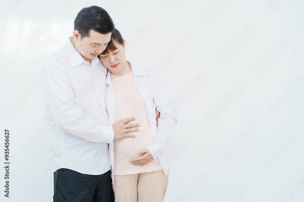 A happy Asian couple, expecting a baby, embraces in a cozy living room. The pregnant woman smiles warmly, while the man lovingly supports her, capturing a moment of joyful anticipation. A happy Asian couple, expecting a baby, embraces in a cozy living room. The pregnant woman smiles warmly, while the man lovingly supports her, capturing a moment of joyful anticipation.