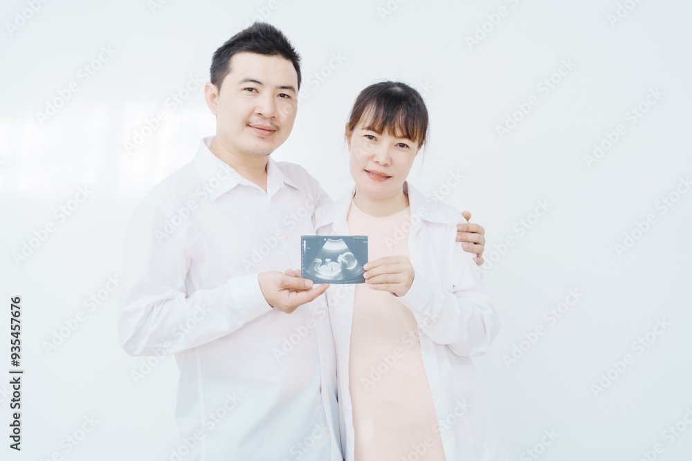A happy Asian couple, expecting a baby, embraces in a cozy living room. The pregnant woman smiles warmly, while the man lovingly supports her, capturing a moment of joyful anticipation. A happy Asian couple, expecting a baby, embraces in a cozy living room. The pregnant woman smiles warmly, while the man lovingly supports her, capturing a moment of joyful anticipation.