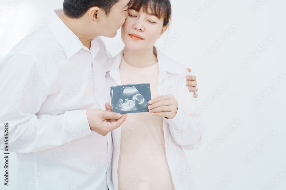 A happy Asian couple, expecting a baby, embraces in a cozy living room. The pregnant woman smiles warmly, while the man lovingly supports her, capturing a moment of joyful anticipation. A happy Asian couple, expecting a baby, embraces in a cozy living room. The pregnant woman smiles warmly, while the man lovingly supports her, capturing a moment of joyful anticipation.