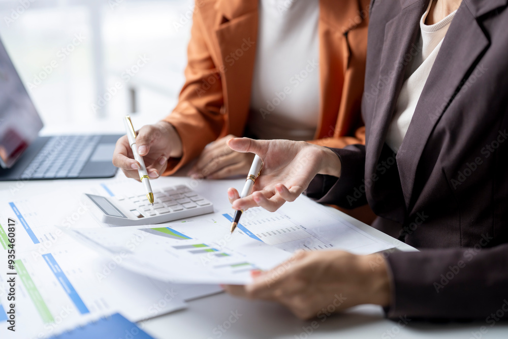 Two businesswomen analyzing financial charts and using calculator in office Two businesswomen analyzing financial charts and using calculator in office
