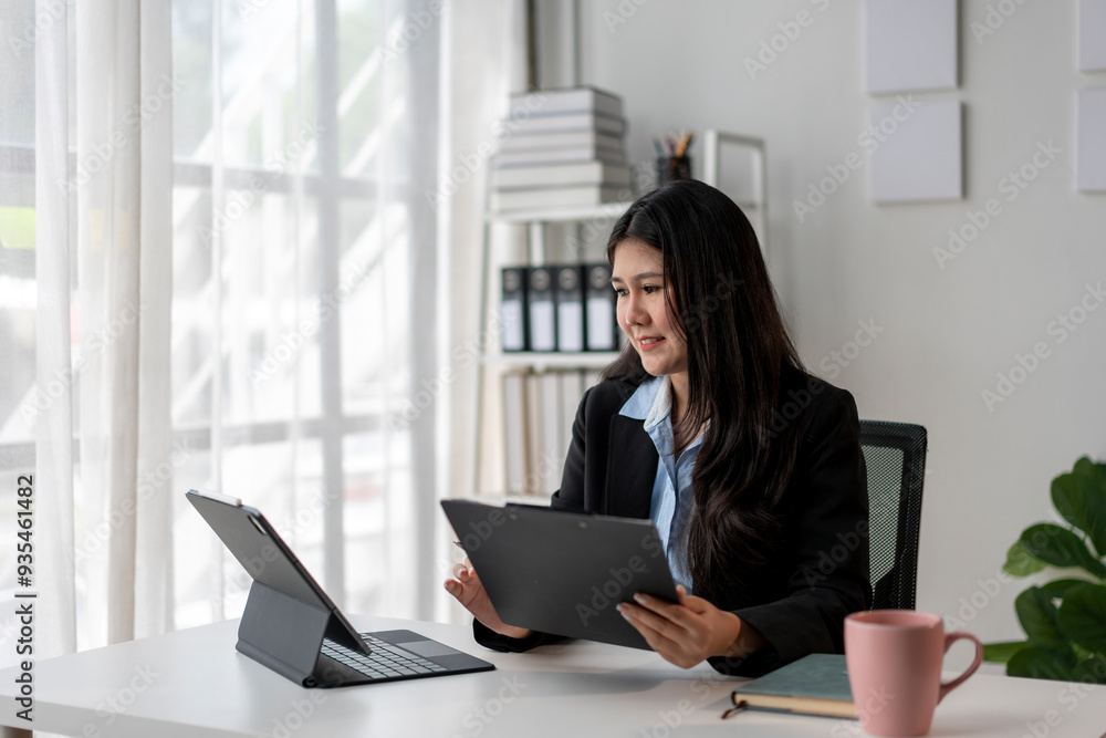 A woman in a business suit is sitting at a desk with two laptops and a clipboard