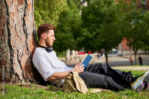 A man is sitting under a tree reading a book. He is wearing headphones and has a backpack next to him. The scene is peaceful and relaxing, with the man enjoying his time outdoors photo