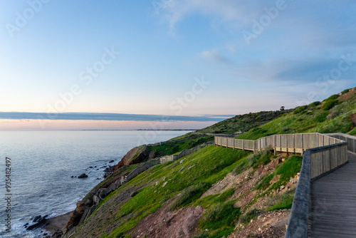 Scenic view of rolling grass hills meeting the ocean, with a charming boardwalk winding through the landscape, capturing the serene beauty of coastal nature.