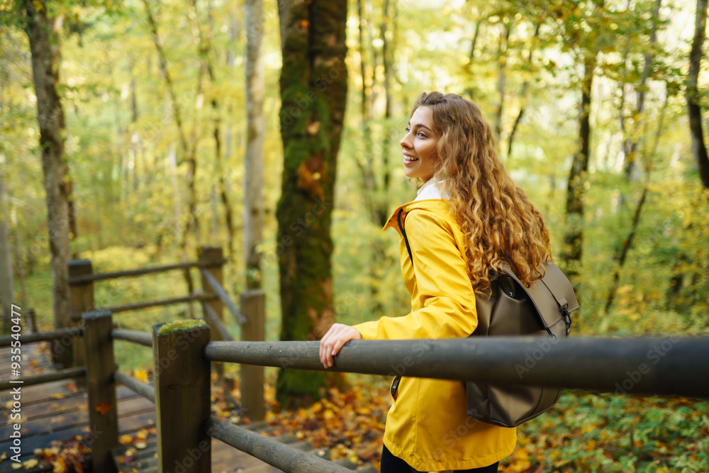 A woman in a yellow raincoat admires nature while walking along a wooden path in a colorful autumn forest A woman in a yellow raincoat admires nature while walking along a wooden path in a colorful autumn forest