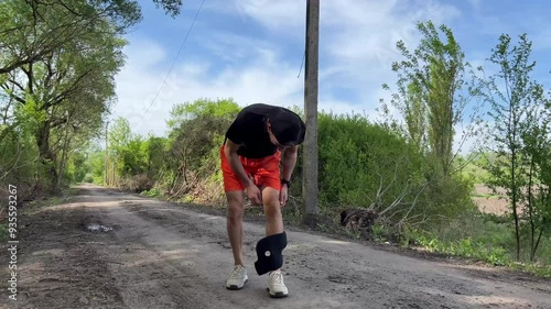 Sportsman in orange shorts take off on a knee support brace while training on a countryside road, promoting injury prevention