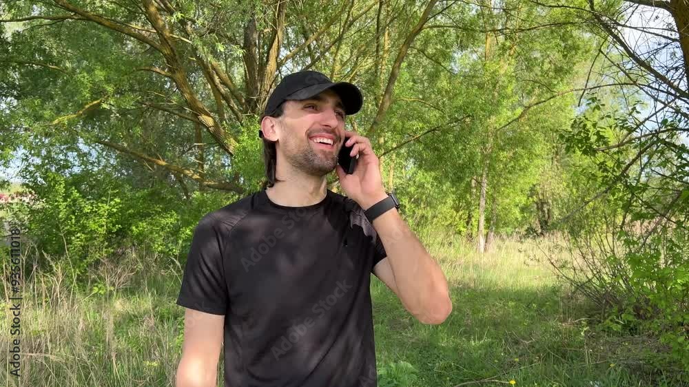 Sportsman standing near a tree, smiling and enjoying a phone call in a vibrant outdoor setting Sportsman standing near a tree, smiling and enjoying a phone call in a vibrant outdoor setting