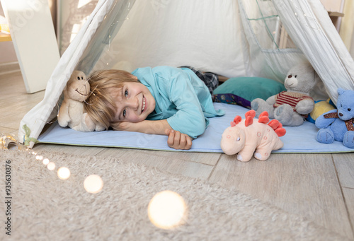 cute little caucasian boy is lying in a wigwam. Childhood, playing with soft toys, tenderness. Children's room with a teepee tent. Cozy atmosphere interior of a children's room