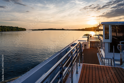 cruiser ship at sunset, chrome railing on sundeck, on river with water, forest, bank in danube delta in romania, with wheel house, romantic with blue sky, blue hour, red sun