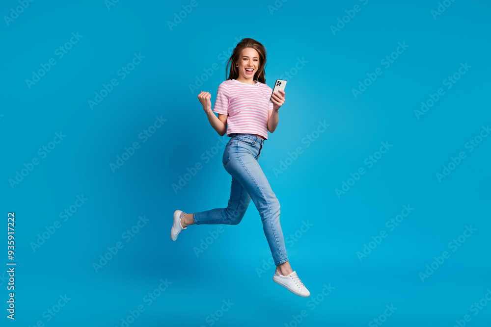 Full body photo of attractive young woman running jump winning hold device dressed striped pink clothes isolated on blue color background Full body photo of attractive young woman running jump winning hold device dressed striped pink clothes isolated on blue color background
