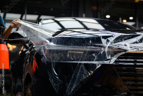 A worker is applying transparent film to a car to protect it from gravel and scratches on the hood of a black car. Applying transparent paint protection film