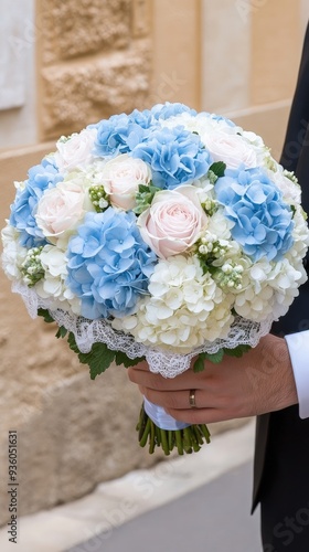 A groom's hand delicately holds a beautifully arranged bouquet of pastel blue and pink flowers against a clean white wall, symbolizing love and celebration photo