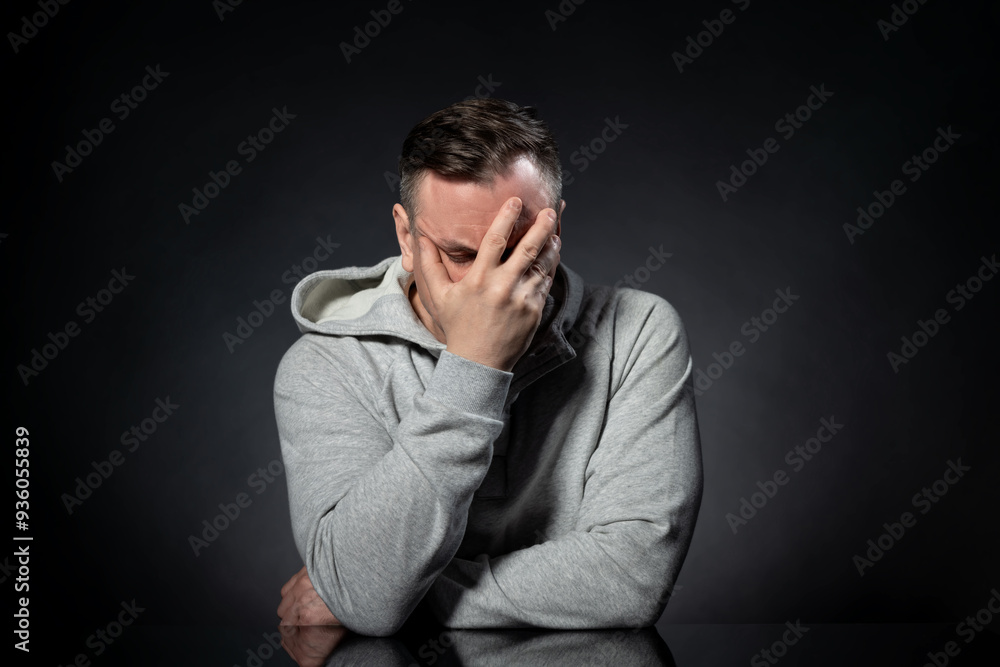 A man in a white hoodie sits at a table and makes a facepalm, on a black background. A man in a white hoodie sits at a table and makes a facepalm, on a black background.