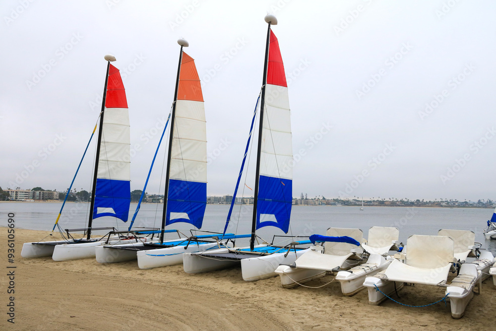 Fototapeta premium Electric and sail catamaran yachts resting on the beach sand under a gloomy sky over Sail Bay in the early morning, San Diego, California