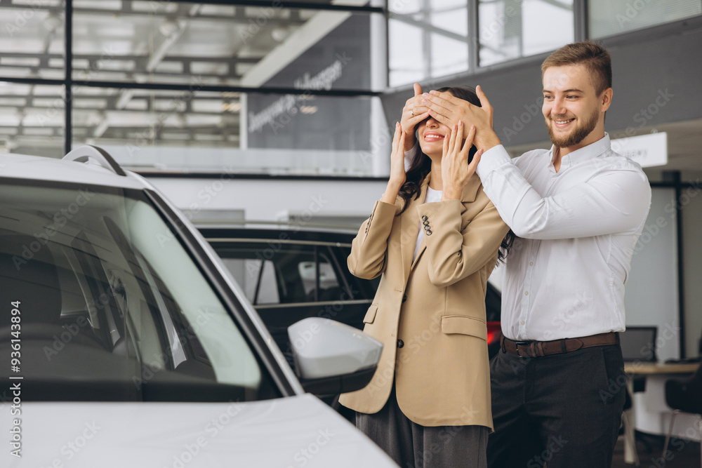 Man close eyes to his wife and makes a surprise buying new car. Happy couple in car showroom.