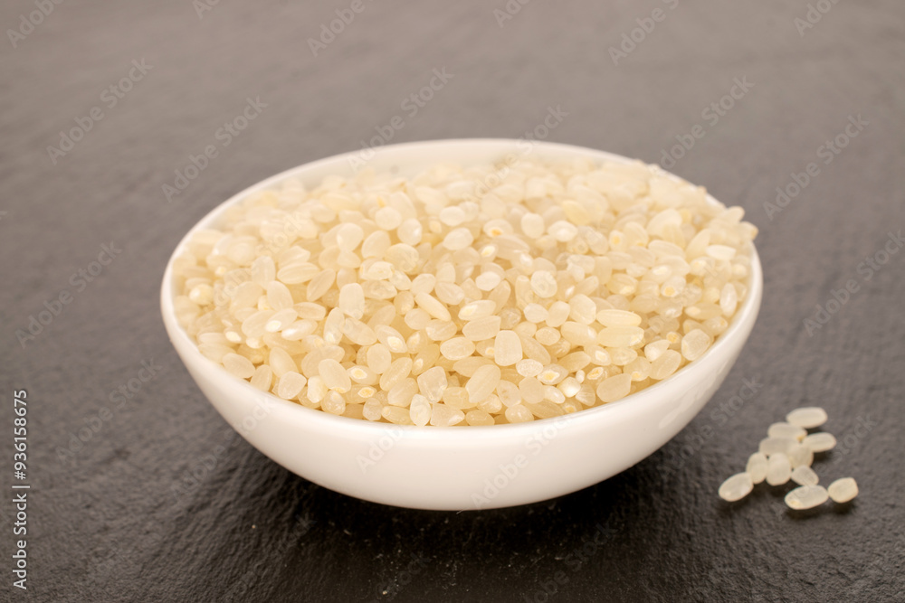 Round grain uncooked rice with white ceramic saucer on slate stone, macro. Round grain uncooked rice with white ceramic saucer on slate stone, macro.