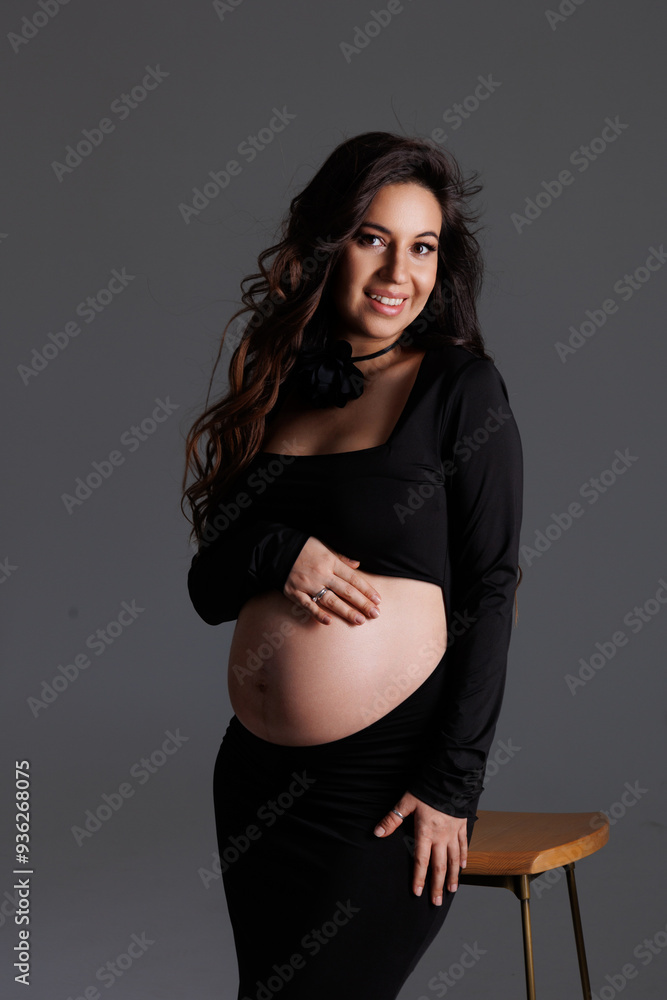 A beautiful pregnant woman with long dark hair with styling and makeup. A brunette waiting for a baby, sit on high chair in a long black dress. Grey background in the studio, fan. A beautiful pregnant woman with long dark hair with styling and makeup. A brunette waiting for a baby, sit on high chair in a long black dress. Grey background in the studio, fan.