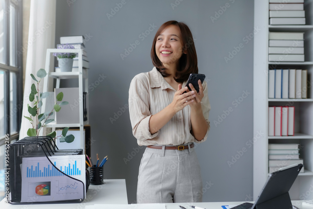 Businesswoman is smiling while using her smartphone in an office with graphs and charts on the desk Businesswoman is smiling while using her smartphone in an office with graphs and charts on the desk