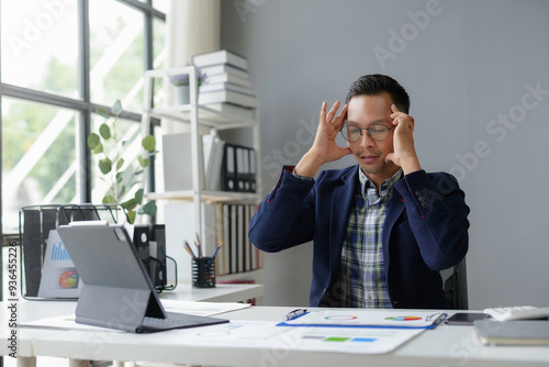 Stressed business man having a headache, touching temples and keeping eyes closed while working with laptop in office