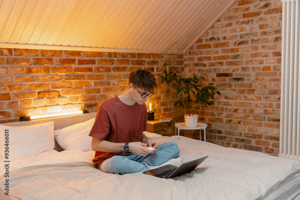 A man in casual clothes uses a laptop while sitting on a bed at home. He puts on his earbuds. Evening time, lamp light A man in casual clothes uses a laptop while sitting on a bed at home. He puts on his earbuds. Evening time, lamp light
