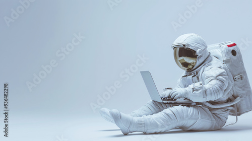 An astronaut in a white suit is seated on the floor, working on a laptop on a clean light gray background with copy space photo