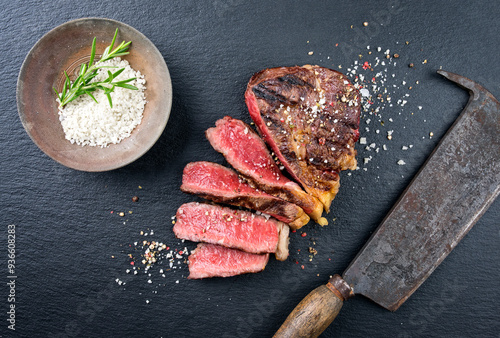 Traditional barbecue dry aged rib-eye beef steak with salt in a bowl sliced as top view on a black design board with a rustic kitchen cleaver photo