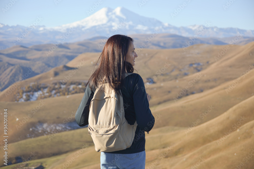 A young brunette woman with a backpack stands against the background of snow-capped Mount Elbrus and looks towards the mountains.