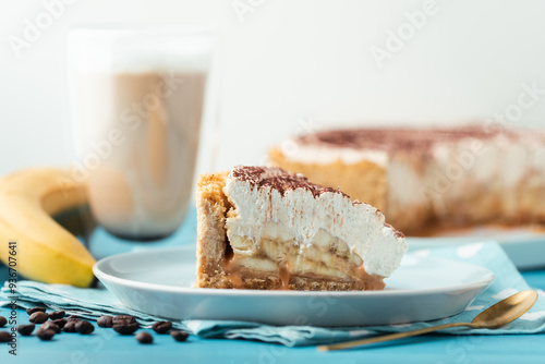 Slice of banoffe pie filled with banana and dulce de leche and sprinkled with grated chocolate. Banana cake on white plate with glass of coffee on turquoise summer background photo