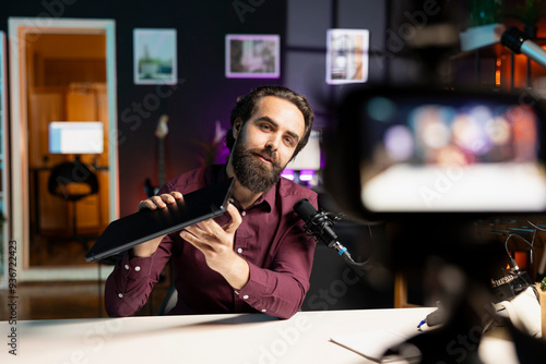 Man presenting laptop to audience, filming video with professional camera and mic. Content creator assessing notebook product, recording with specialized equipment in studio photo